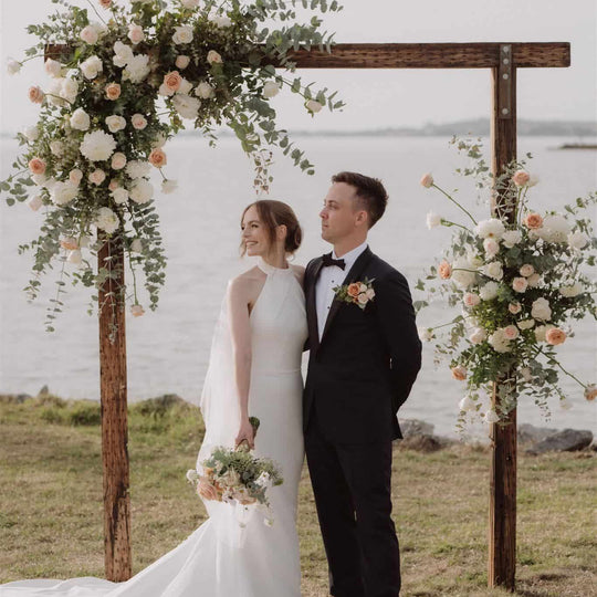 Wedding couple standing under a floral arch by a body of water.
