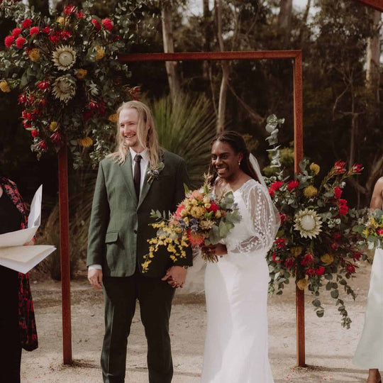 Wedding couple standing under a floral archway with a natural outdoor setting.